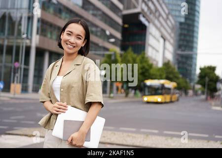 Schöne asiatische Mädchen lächelt als pendelt zur Arbeit, steht auf der Straße mit Laptop und Notebook Stockfoto