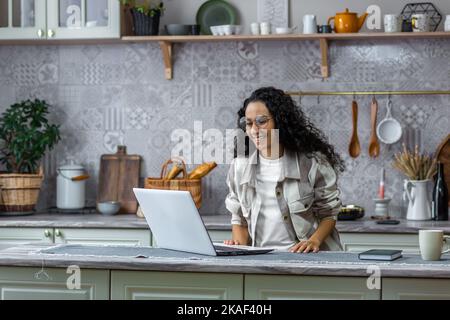 Glückliche hispanische Frau zu Hause, die lächelnd auf den Laptop schaut und aus der Ferne mit Freunden spricht. Stockfoto