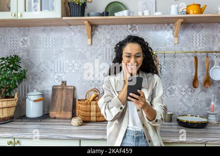 Glückliche lateinamerikanische Frau zu Hause, die gute Online-Nachrichten vom Smartphone liest, Frau in Brille und lockiges Haar, die mit dem Telefon Online-Inhalte zu Hause in der Küche anschaut. Stockfoto