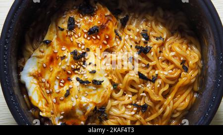 Eine Nahaufnahme von oben aus gekochten Instant-Nudeln mit einem Ei und Sesam in einer dunklen Schüssel Stockfoto