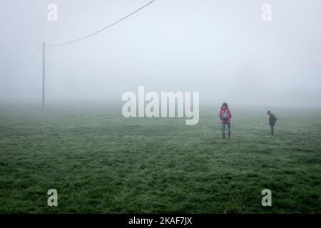 Ein junges Mädchen und ihr kleiner Bruder, die mit ihren Rucksäcken auf einem Feld im Nebel herumlaufen. Stockfoto