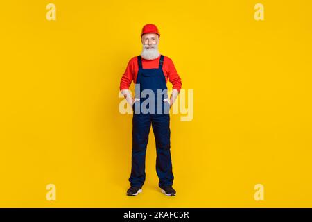 In voller Länge Foto von guten Laune Senior Mann Baumeister mit weißem Bart roten Helm Hände in Taschen isoliert auf gelbem Hintergrund Stockfoto