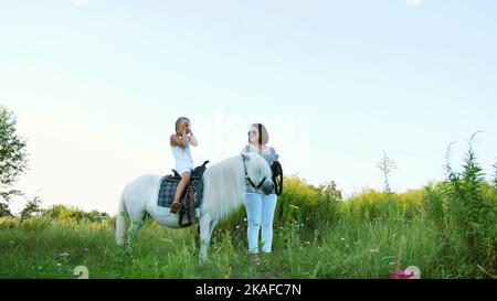 Mutter und Tochter laufen auf dem Feld herum, Tochter reitet auf einem Pony, Mutter hält ein Pony für einen Zaum. Fröhlicher, glücklicher Familienurlaub. Im Freien, im Sommer, in der Nähe des Waldes. Hochwertige Fotos Stockfoto