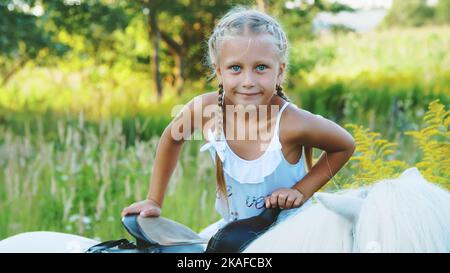 Blonde Mädchen von sieben Jahren, in einem weißen Kleid, klettert, sitzt auf einem Pony. Fröhlicher, glücklicher Familienurlaub. Im Freien, im Sommer, in der Nähe des Waldes. Hochwertige Fotos Stockfoto