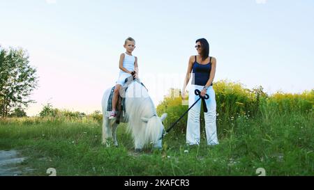 Eine Frau und ein Junge gehen um das Feld, Sohn reitet auf einem Pony, Mutter hält ein Pony für einen Zaum. Fröhlicher, glücklicher Familienurlaub. Im Freien, im Sommer, in der Nähe des Waldes. Hochwertige Fotos Stockfoto