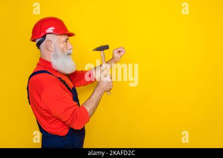 Foto von ernsthaften professionellen alten Kerl gekleidet Uniform insgesamt roten Hardhat Hummer in Nägeln leeren Raum isoliert gelben Hintergrund Stockfoto