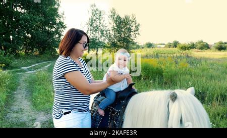 Eine Frau mit einem Baby geht um das Feld, ein Baby sitzt auf einem Pony, Mama hält das Baby. Fröhlicher, glücklicher Familienurlaub. Im Freien, im Sommer, in der Nähe des Waldes. Hochwertige Fotos Stockfoto