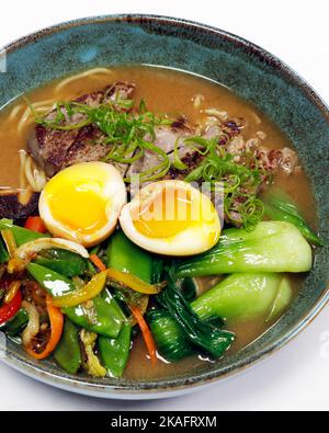 asiatische Spezialität Ramen Nudelsuppe mit Rinderstreifen, pochierten Eiern, Bok Choi und Sommergemüse in einer Schüssel Stockfoto