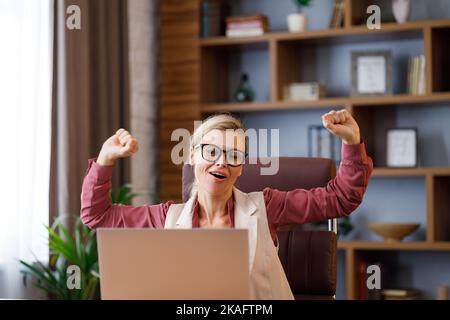 Glückliche Geschäftsfrau überrascht von den guten Nachrichten beim Blick auf Laptop sitzen im Büro.Manager genießen Erfolg, feiern Sieg, machen gute Geschäfte Stockfoto