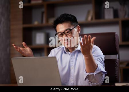 Ein asiatischer Mann hält ein Online-Webinar mit einer Laptop-Webcam ab und sitzt in einem Heimbüro. Mann, der Online-Meetings hat und ferngesteuert kommuniziert, schaut sich an Stockfoto