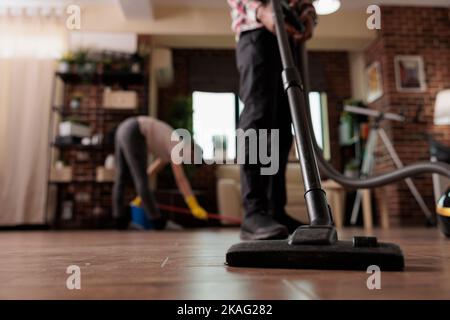 Mann saugt Schmutz vom Boden, während Frau Latexhandschuhe und Mops trägt. Multirassisches Paar, das Hausarbeit am freien Tag von der Arbeit tut und zusammen arbeitet, um ein harmonisches Zuhause zu haben. Stockfoto