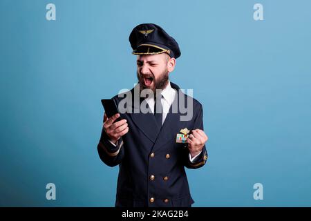 Glücklich lächelnder Flugzeugpilot in Uniform hält modernes Telefon mit Gewinnergeste, geballte Faust. Pilot mit fröhlichem Gesichtsausdruck, der auf den Bildschirm des Mobiltelefons schaut und gute Nachrichten liest Stockfoto