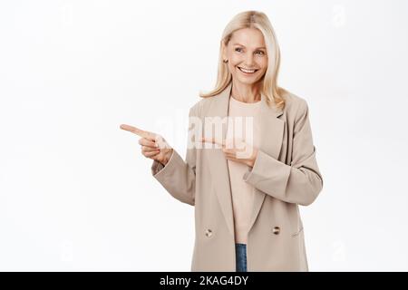 Porträt einer lächelnden blonden, reifen Frau, Dame, die glücklich aussieht, mit dem Finger nach links zeigt, Werbung zeigt, die auf weißem Hintergrund steht. Stockfoto