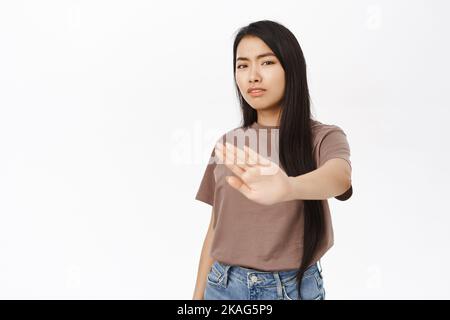 Halt, bleibe zurück. Besorgte asiatische Frau streckt ihre Hand, um zu verbieten, smth abzulehnen, Aktion zu verbieten, Niedergang smth schlecht, steht über weißem Hintergrund. Stockfoto