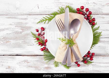 Weihnachts- oder Wintergeschirr. Besteck und Serviette auf einem weißen Teller mit Rahmen aus immergrünen Ästen und roten Beeren. Oben Blick auf einen weißen Holzhintergr Stockfoto