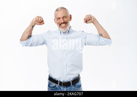 Starrköpfiger und selbstbewusster reifer Mann mit grauem Haar, lächelnd und biegsam Bizeps, zeigt Muskeln mit fröhlichem Gesicht, steht auf weißem Hintergrund Stockfoto