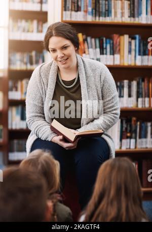 Sie liebt es, alle Fragen über die Geschichte zu beantworten. Eine Grundschullehrerin liest ihren Kindern in der Bibliothek vor. Stockfoto