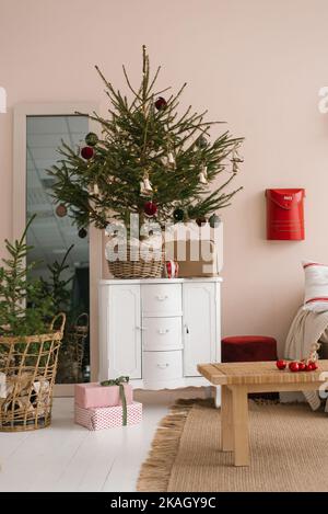 Stilvoller Weihnachtsbaum mit Spielzeug im Korbkorb steht auf einer weißen Kommode im Wohnzimmer Stockfoto