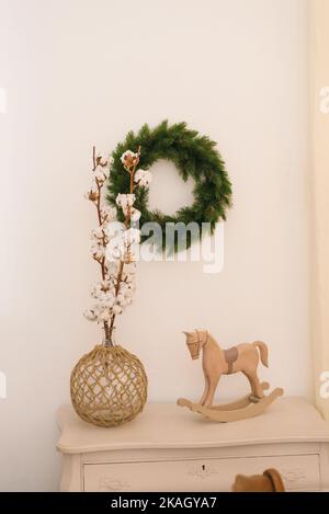Baumwollzweige in einer Vase, ein Spielzeug-Holzpferd auf der Kommode, ein Weihnachtskranz an der Wand Stockfoto