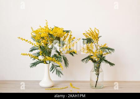 Wunderschönes Frühlingsbouquet aus gelben Mimosenzweigen in Keramikvase und Glasflasche, das auf einem Holzregal vor weißem Hintergrund steht. Stockfoto