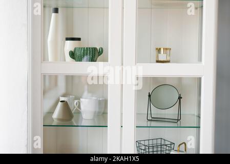 Dekorative Gegenstände auf den Regalen im Schrank hinter dem Glas im Inneren des Wohnzimmers. Vasen, Tischspiegel, Kerze, Tassen Stockfoto