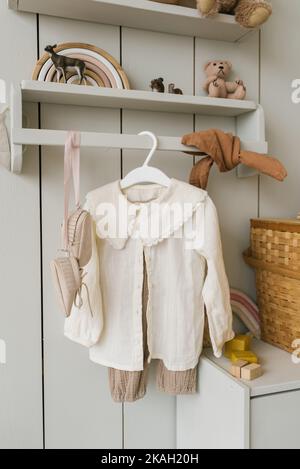 An einem Kleiderbügel im Kinderzimmer hängen Kinderkleidung und verschiedene Spielzeuge im minimalistischen Stil Stockfoto