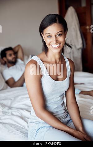 Steh auf und glänze zu einem großartigen Tag. Porträt einer jungen Frau, die zu Hause auf einem Bett mit ihrem Mann im Hintergrund sitzt. Stockfoto