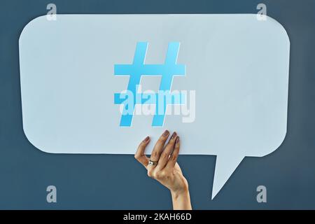 Sag es mit einem Hashtag. Beschnittene Studioaufnahme einer Frau, die eine Sprechblase mit einem Hashtag auf einem grauen Hintergrund hochhält. Stockfoto