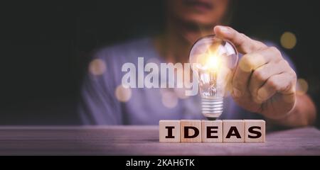 Hand berühren auf Glühbirne auf Holzblock mit Word-Ideen, neues Ideenkonzept mit Innovation und Inspiration, innovative Technologie in Wissenschaft und Kommunikation Stockfoto