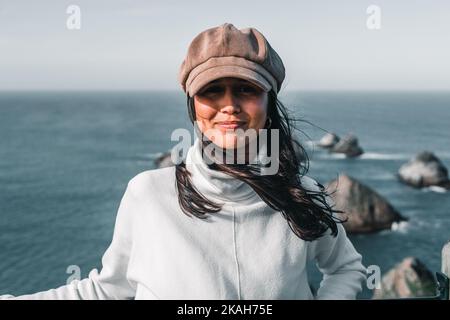 kaukasisches Mädchen mit weißem Pullover und langen schwarzen Haaren braunen Cord Hut Blick auf Kamera glücklich aus Sicht über Felsen und Meer, Nugget Point, neu Stockfoto