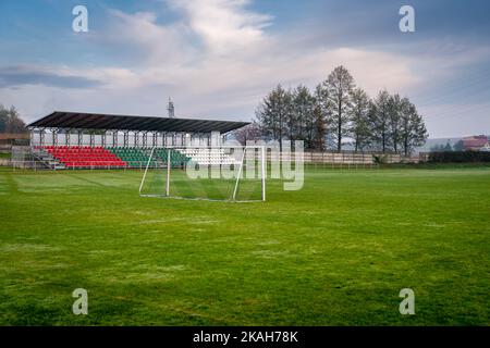 Lokales Fußballstadion mit kleinen Sport-Tribüne durch das Tor gesehen. Bäume im Hintergrund. Herbsttag auf dem Land. Stockfoto