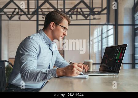 Ein vielbeschäftigter Mann mit Brille, der in seinem Büro online am Laptop arbeitet, ein Finanzexperte, der ein Diagramm auf dem Bildschirm des Monitors mit Diagramm analysiert und in einem Notizblock schreibt. Kryptowährung Stockfoto