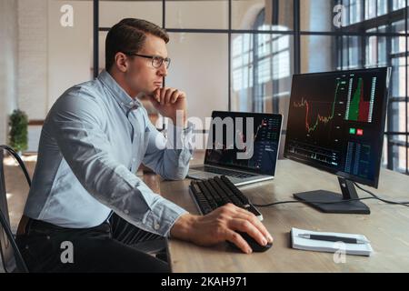 Seitenansicht des Krypto-Brokers, der am Bürotisch sitzt, den Markt analysiert, den Bildschirm des PCs mit einem Candlestick-Chart betrachtet, das Kinn mit der Hand berührt, denkt, Hemd und Brille trägt Stockfoto