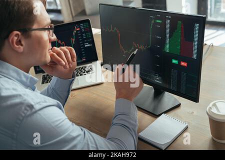 Konzentrierter Geschäftsmann, der auf einen pc-Monitor mit Diagramm des Marktes für Krypto-Aktien schaut, mit Stift auf den Bildschirm zeigt, das Kinn berührt, die Tendenz des Geldflusses analysiert und eine neue Strategie erfindet Stockfoto