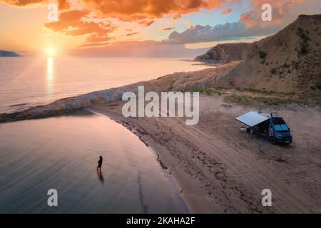 Luftaufnahme des Wohnwagens am verlassenen Strand vor einem wunderschönen Sonnenuntergang. Menschen, die im glitzernden Meer baden. Outdoor Nomaden Lifestyle. Stockfoto