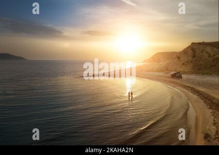 Luftaufnahme des Wohnwagens am verlassenen Strand vor einem wunderschönen Sonnenuntergang. Menschen, die im glitzernden Meer baden. Outdoor Nomaden Lifestyle. Stockfoto