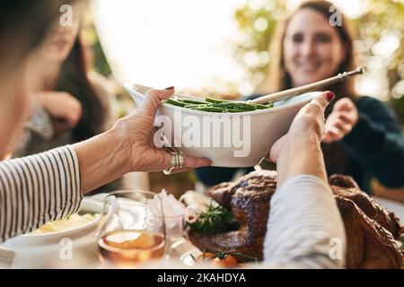 Bitte übergeben Sie die Bohnen. Eine unkenntliche Frau, die mittags über einen Tisch eine Platte mit grünen Bohnen an eine fröhliche Frau übergibt. Stockfoto