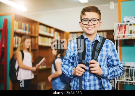 Ich bin das neue Kind in der Schule. Porträt eines fröhlichen jungen Jungen, der tagsüber in einer Schultasche steht. Stockfoto