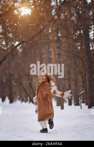 Ein weiß blondes Mädchen in einem Strickhut, einem langen Schal und einem Pelzmantel geht durch den Wald und lächelt. Frau fängt Schnee in weißen Fäustlingen. Der Winter ist auf der Straße. Hochwertige Fotos. Stockfoto