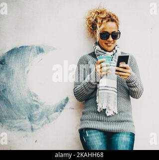 Porträt einer jungen erwachsenen Frau mit einem modernen Mobiltelefon, das an einer weißen Wand mit blauem Mond steht. Konzept von Menschen, die Online-Aktivitäten mit genießen Stockfoto