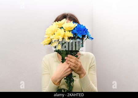 Mädchen hält patriotische blaue und gelbe Blumen vor ihrem Gesicht auf hellem Hintergrund. Gemalte Chrysanthemen als Staatsflagge der Ukraine. Speicherplatz kopieren Stockfoto