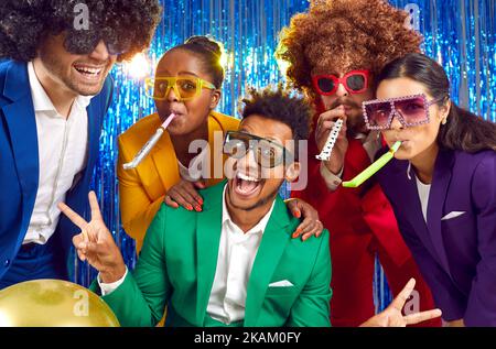 Fröhliche Menschen in bunten Kostümen, witzigen Disco-Brillen und Perücken, die gemeinsam Party-Hörner blasen Stockfoto