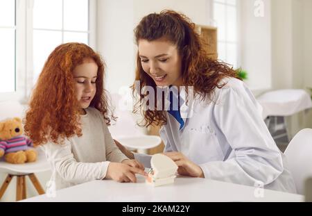 Die Zahnärztin erzählt dem kleinen Mädchen von der Mundhygiene, indem sie ihren künstlichen Kiefer mit Zähnen zeigt. Stockfoto