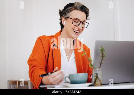 Lächelnde Geschäftsfrau, die an einem virtuellen Meeting teilnimmt, während sie in einem Café arbeitet. Eine glückliche, reife Geschäftsfrau, die während der Arbeit einen Videoanruf mit ihrem Team führt Stockfoto