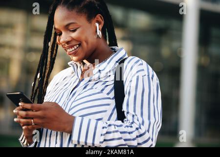 Fröhliche junge Geschäftsfrau, die auf ihrem Weg zur Arbeit in der Stadt ein Smartphone benutzt. Glückliche junge Geschäftsfrau, die während des Pendelns Musik am Telefon spielt Stockfoto