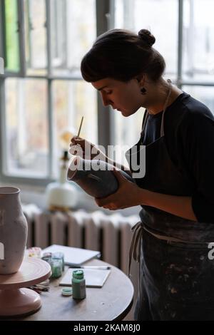 Emotionslose Frau Brünette in Schürze arbeitet als Künstlerin in Keramiker Shop steht neben Tisch mit Werkzeugen Stockfoto