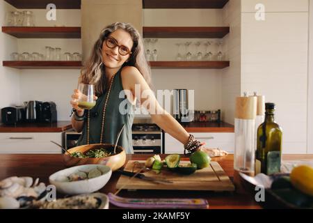 Vegetarierin lächelt an der Kamera, während sie etwas grünen Saft in der Hand hält. Reife Frau, die sich gesunde pflanzliche Nahrung in ihrer Küche serviert. Glücklich sen Stockfoto
