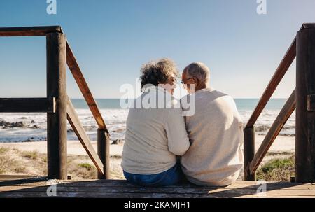 Ein glückliches Ehepaar lächelt sich gegenseitig an, während sie auf einer hölzernen Fußbrücke vor dem Meer sitzt. Ältere Rentner paar Ausgaben einige Qualität ti Stockfoto