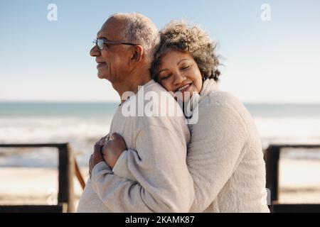 Liebevolle ältere Frau, die fröhlich lächelt, während sie ihren Mann am Meer umarmt. Ein romantisches, älteres Paar genießt es, gemeinsam etwas Zeit zu verbringen Stockfoto