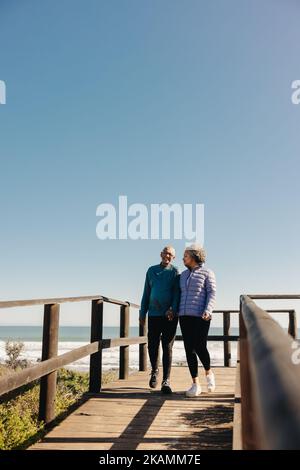 Romantisches Seniorenpaar, das fröhlich lächelt, während es gemeinsam auf einer Fußgängerbrücke am Meer entlang geht. Rentnerehepaar verbringt eine gute Zeit miteinander Stockfoto